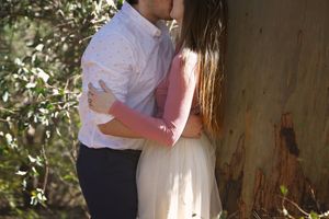 Couple in white shirt and flowing cream skirt share a kiss beside a eucalyptus tree trunk in a sunlit outdoor engagement photo.