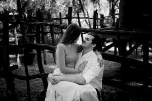 Black and white portrait of a woman sitting on a man's lap on a rustic wooden bench in a romantic outdoor couple photo shoot.