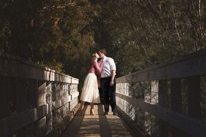Couple in casual formal attire kiss on a wooden bridge framed by trees in a romantic outdoor engagement photography session.