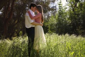 Couple embraces in a tall green grass field surrounded by trees in warm sunlight for a romantic engagement photo.