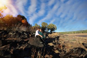 Couple sits close together on dark volcanic rocks under a bright blue sky with wispy clouds and sun flare.