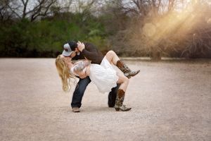 Man dips a woman in a romantic kiss on a gravel path, she wears a white dress and cowboy boots at sunset.