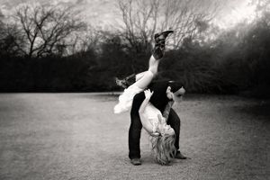 Black and white photo of a playful couple outdoors, the woman flipping upside down while the man in jeans holds her near bare trees.