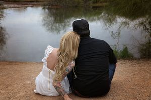 Couple in an engagement session sits at the edge of a calm pond, the blonde woman in a white lace dress leaning on a man in a black cap.