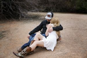 Man in a black cap and woman in a white dress and cowboy boots embrace while sitting on a dirt trail in a western-style couple photo shoot.