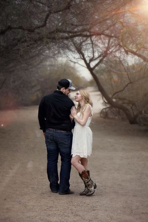 Couple stands close on a desert trail at sunset, the woman in a white dress and cowboy boots gazing up at the man in a cap.
