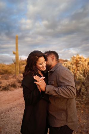 Smiling woman shows her engagement ring as a man kisses her cheek, with a saguaro cactus and cloudy desert sky behind them.