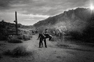 Black and white desert engagement photo of a man dipping his partner for a kiss, with saguaro cacti and rugged mountains behind them.