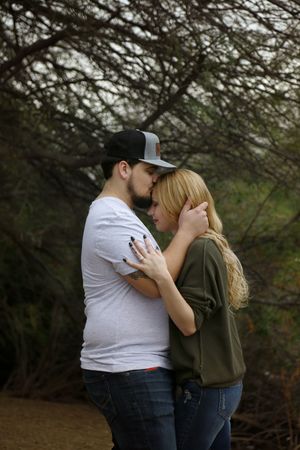 Bearded man in a gray cap kisses the forehead of a blonde woman in an olive sweater during an engagement photo session among bare branches.