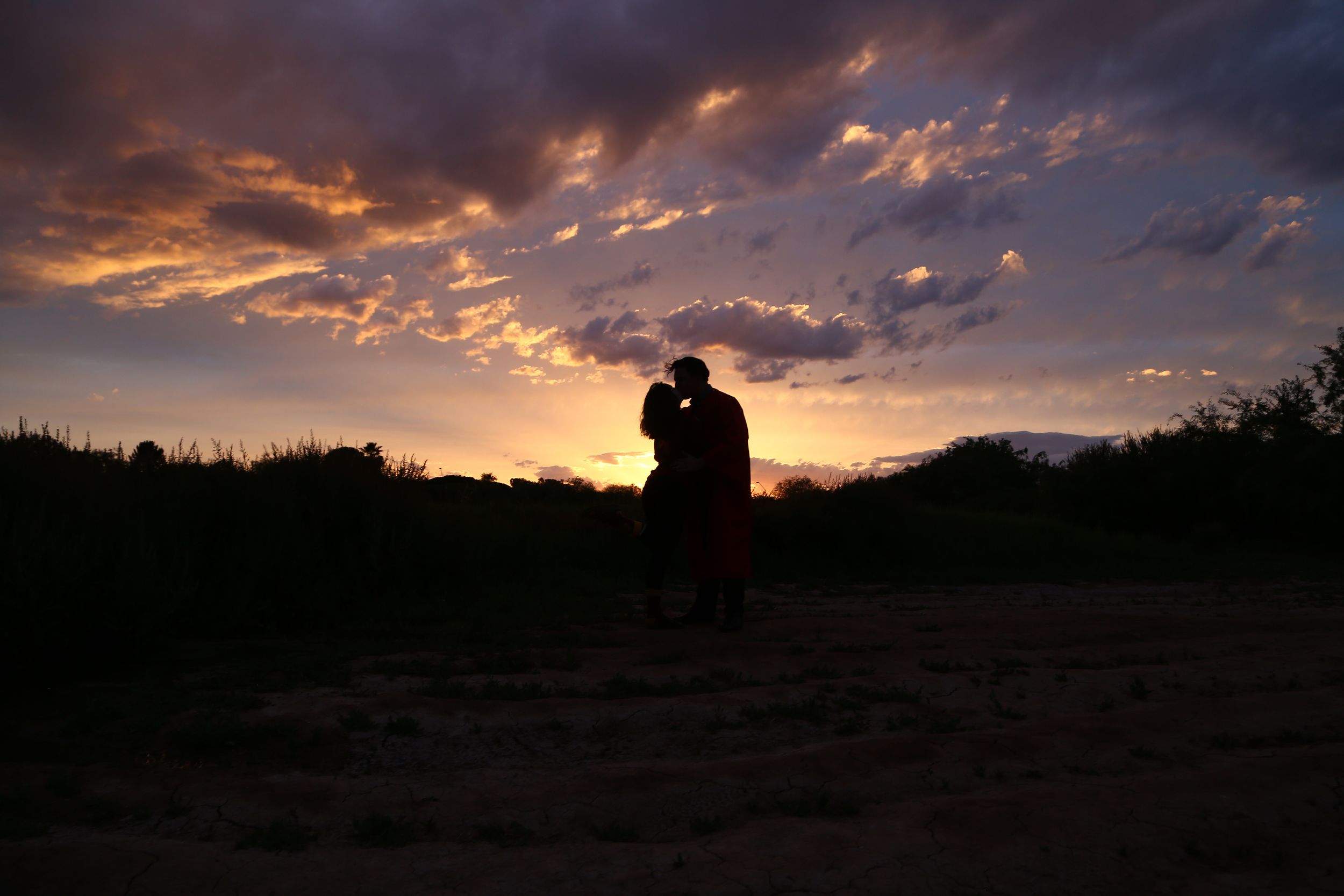 Silhouette of a couple kissing against a dramatic orange and purple sunset sky over a dark desert landscape.