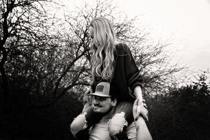Black and white photo of a smiling man in a cap carrying a long-haired blonde woman on his shoulders among bare trees.