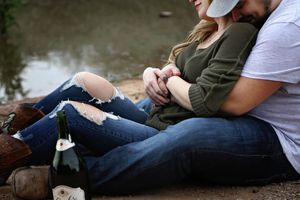 Couple in jeans and boots sitting close beside a pond, holding hands, with a champagne bottle resting nearby.