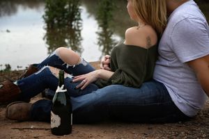 Couple cuddling by a reflective pond with a champagne bottle in the foreground during an outdoor engagement session.