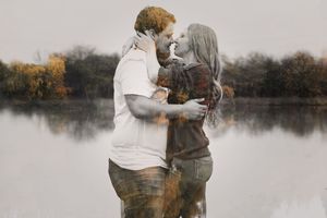 Couple sharing a romantic kiss beside a calm lake with autumn trees in a soft, dreamy double-exposure style photo.