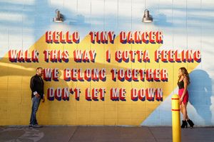 Couple standing apart in front of a yellow brick wall painted with colorful lyrics about dancing and belonging together.
