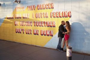Couple embracing and kissing beside a mural wall featuring song lyrics in red lettering on a yellow and white background.