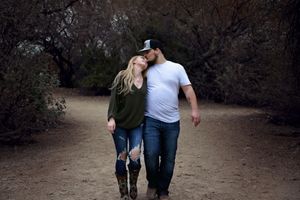 Couple walking together along a wooded dirt trail, embracing and sharing a tender forehead kiss in casual outfits.