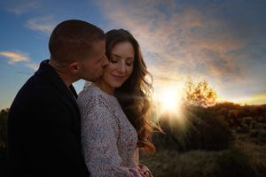 Couple cuddling at sunset as a man kisses the cheek of a smiling woman with long brown hair against a glowing sky.