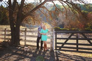 Engaged couple standing close beside a rustic wooden ranch fence under a large oak tree, with a small dog at their feet.