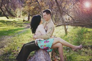 A man in a military uniform holds a dark-haired woman in a floral skirt as she sits on a log in a sunny park.