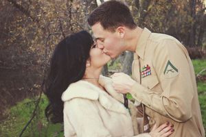 A uniformed service member kisses a dark-haired woman in a cream fur coat among autumn trees in a romantic outdoor portrait.