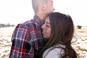 Woman with windblown hair nuzzles a man in a red plaid flannel shirt during a winter couple portrait session outdoors.