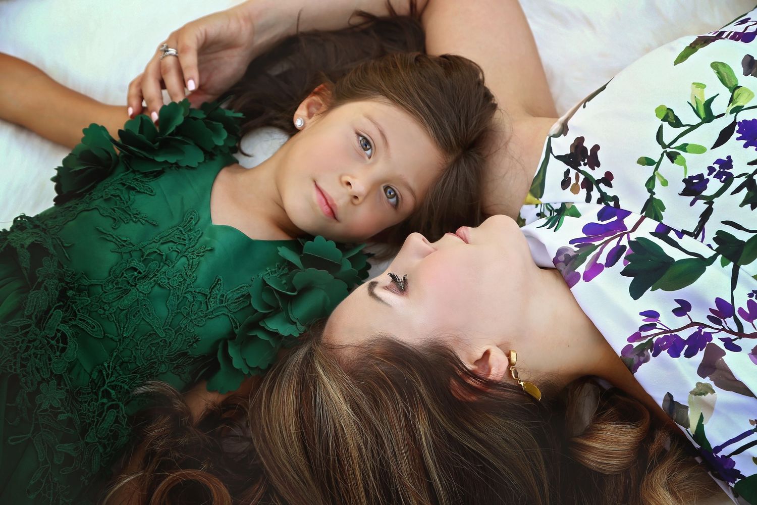 A mother in a floral top lies close to her young daughter in a green ruffled dress, resting together on a soft white bed.