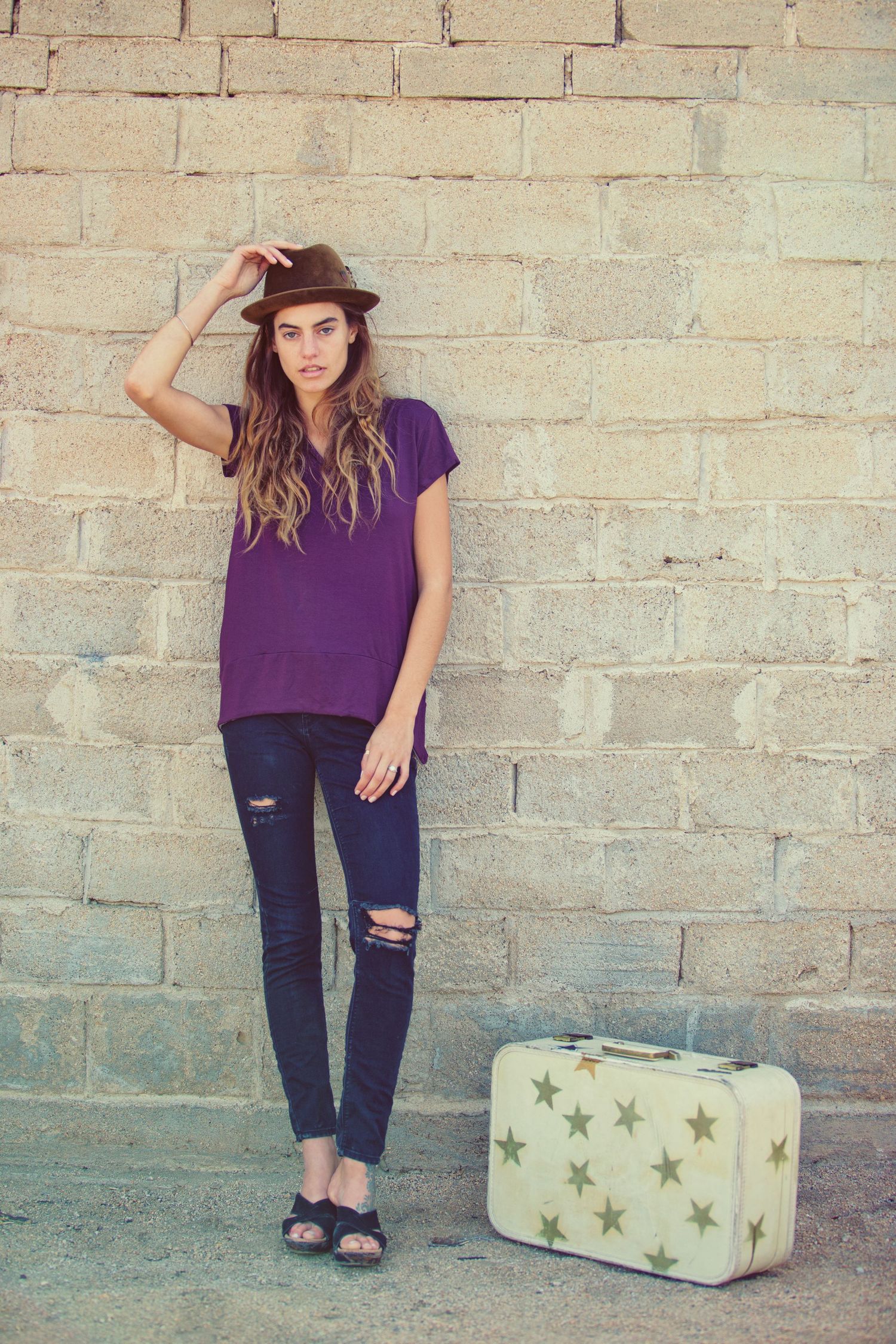 Young woman in purple top, ripped jeans, and brown fedora poses against stone wall with vintage star-print suitcase.