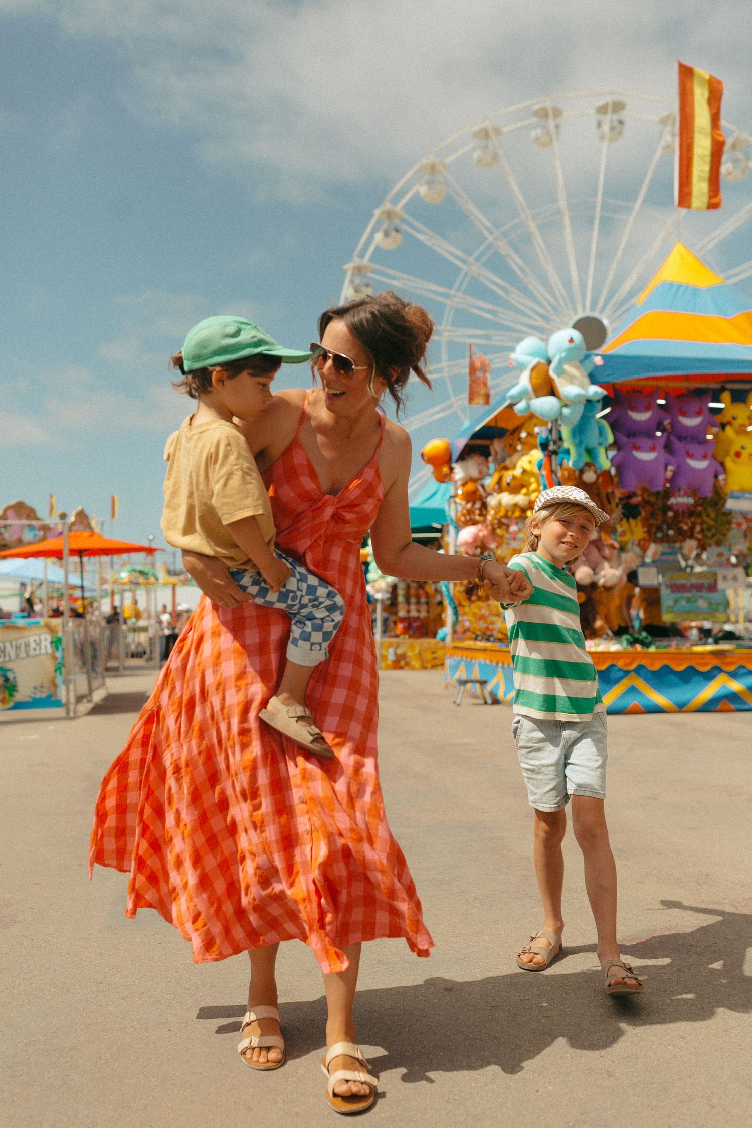 A mother in an orange plaid dress carries her young son at a colorful summer fair with a Ferris wheel backdrop.