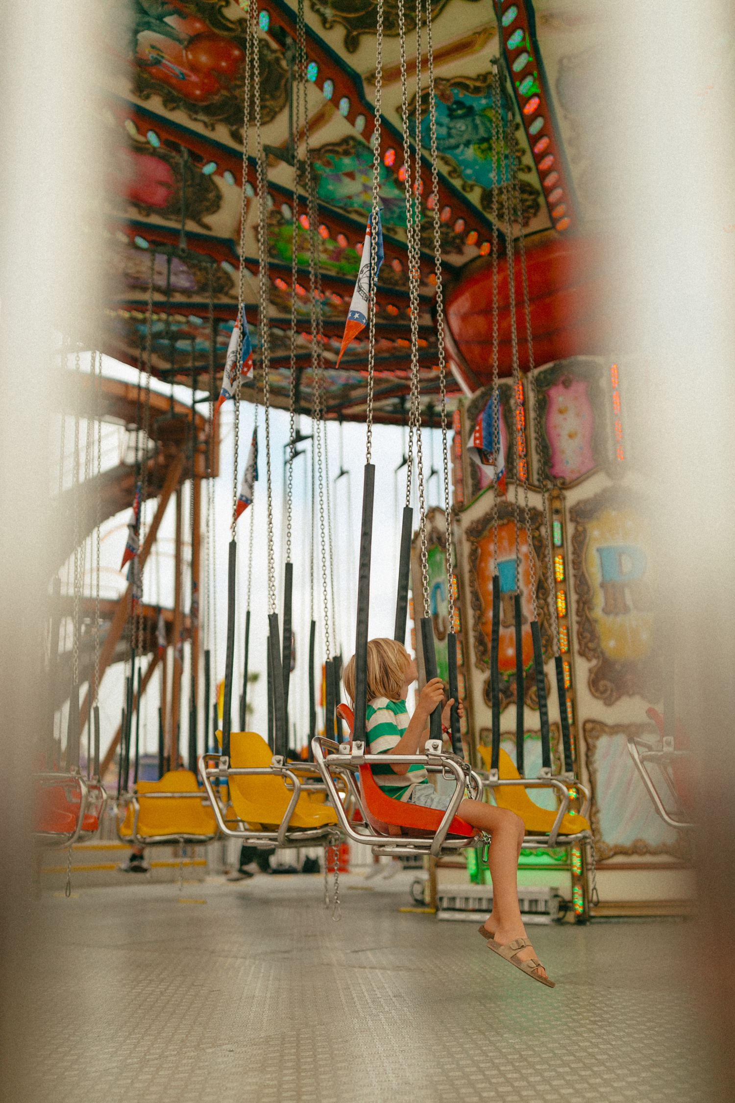 A child sits on a colorful chain swing carousel ride at an amusement park, surrounded by vibrant decorations.