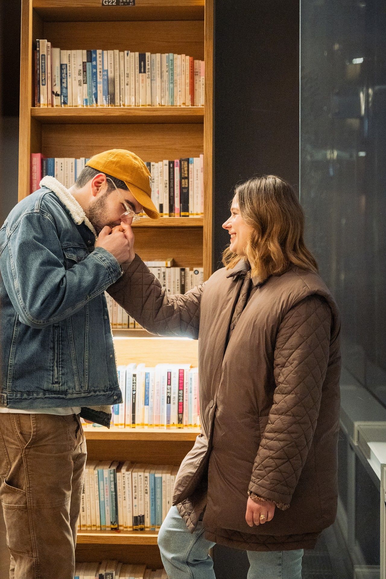 A man in a denim jacket kisses a woman's hand near a wooden bookshelf in a cozy library setting.