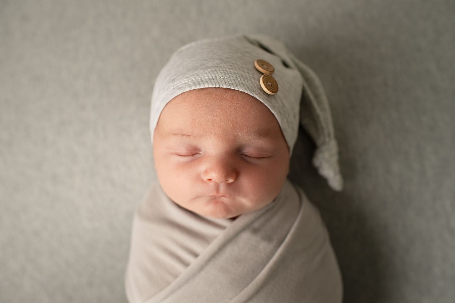 Sleeping newborn baby wrapped in gray swaddle wearing a knot hat with buttons.