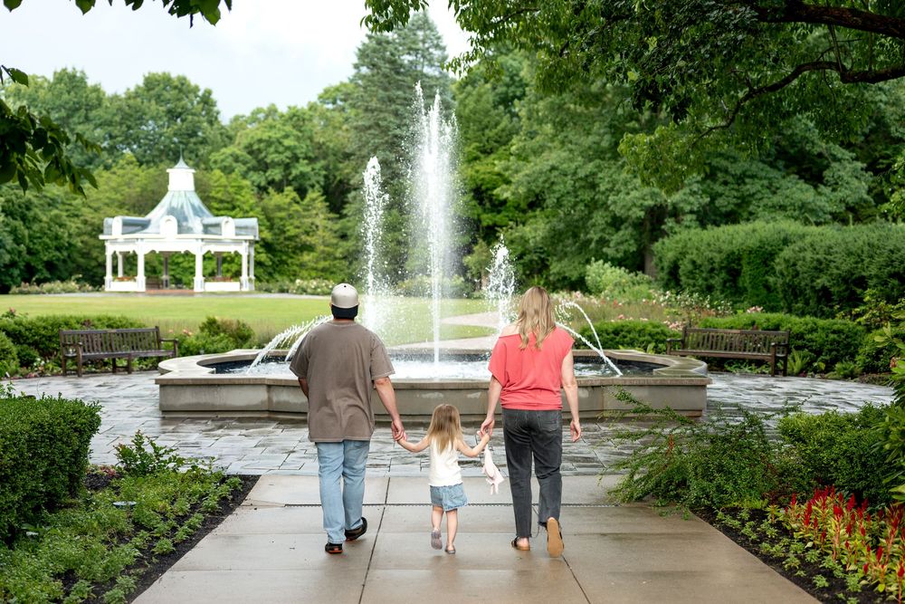 Family of three holding hands walking toward a fountain in a lush green park with a white gazebo.