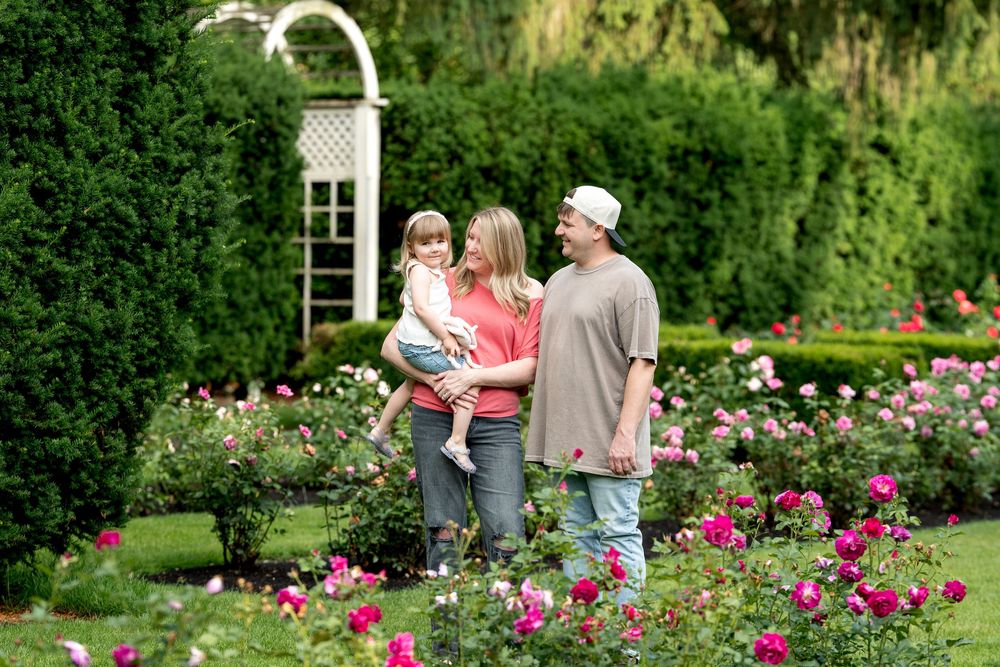 Family with young child posing among blooming pink roses in a lush green garden with an arbor.