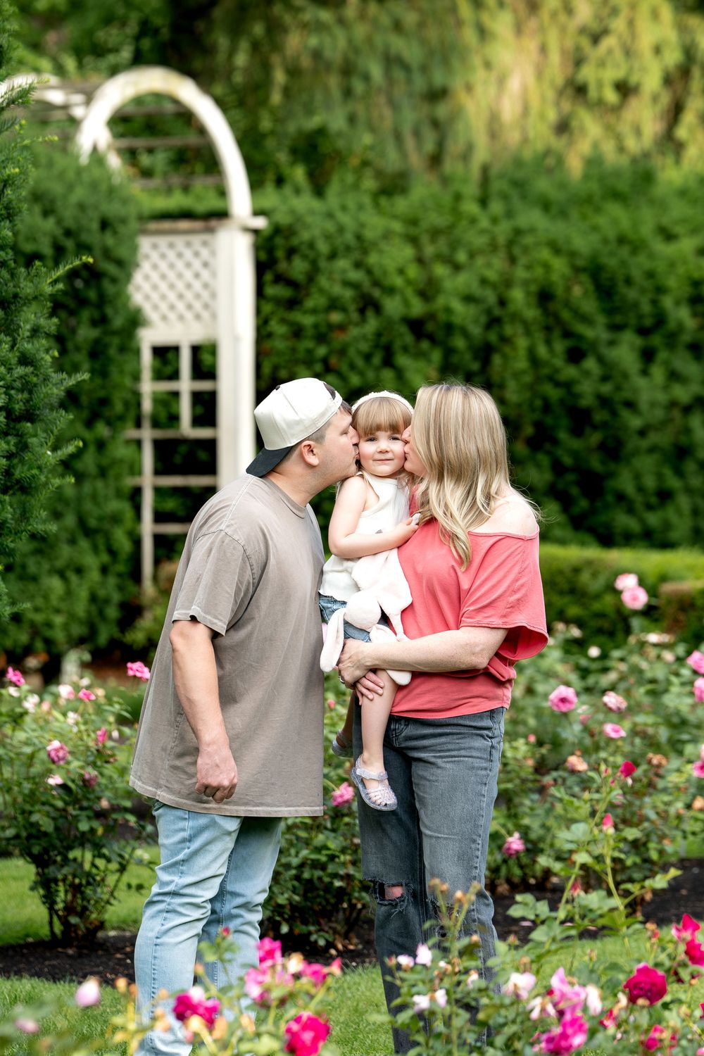 Parents kissing their young daughter among blooming roses in a garden.