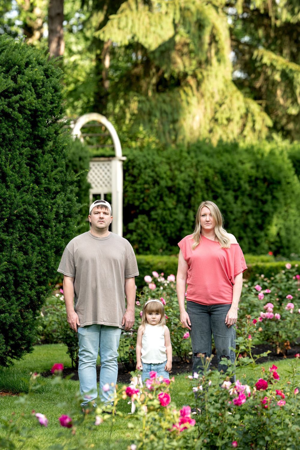 Family of three posing in a lush garden with blooming roses and tall green trees in the background.