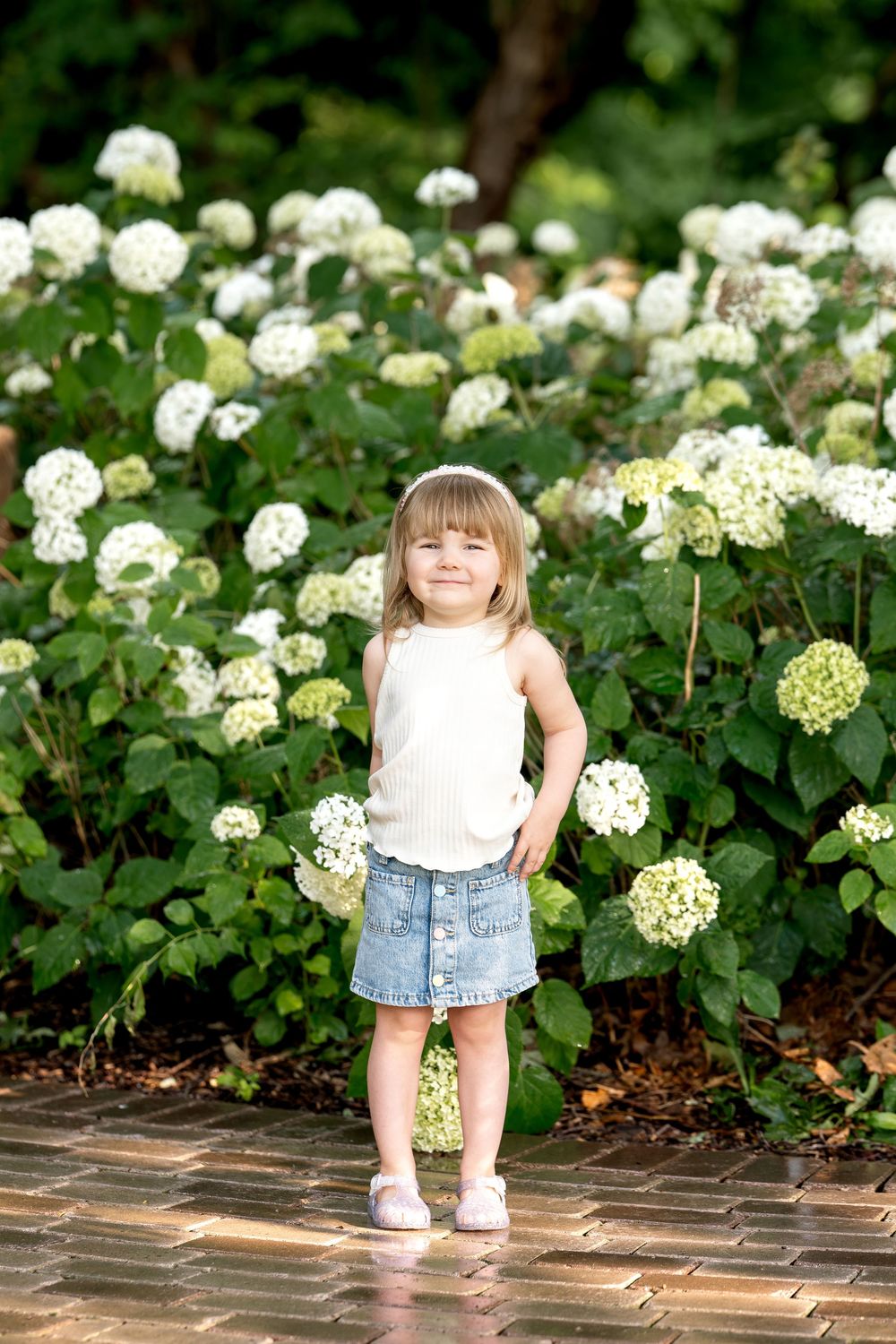 Young girl in white top and denim skirt smiling in front of blooming white hydrangea bushes outdoors.