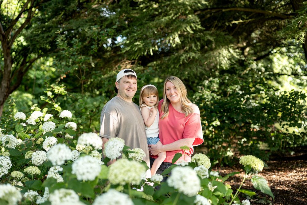 Family of three smiling among white hydrangeas in a lush green garden setting.