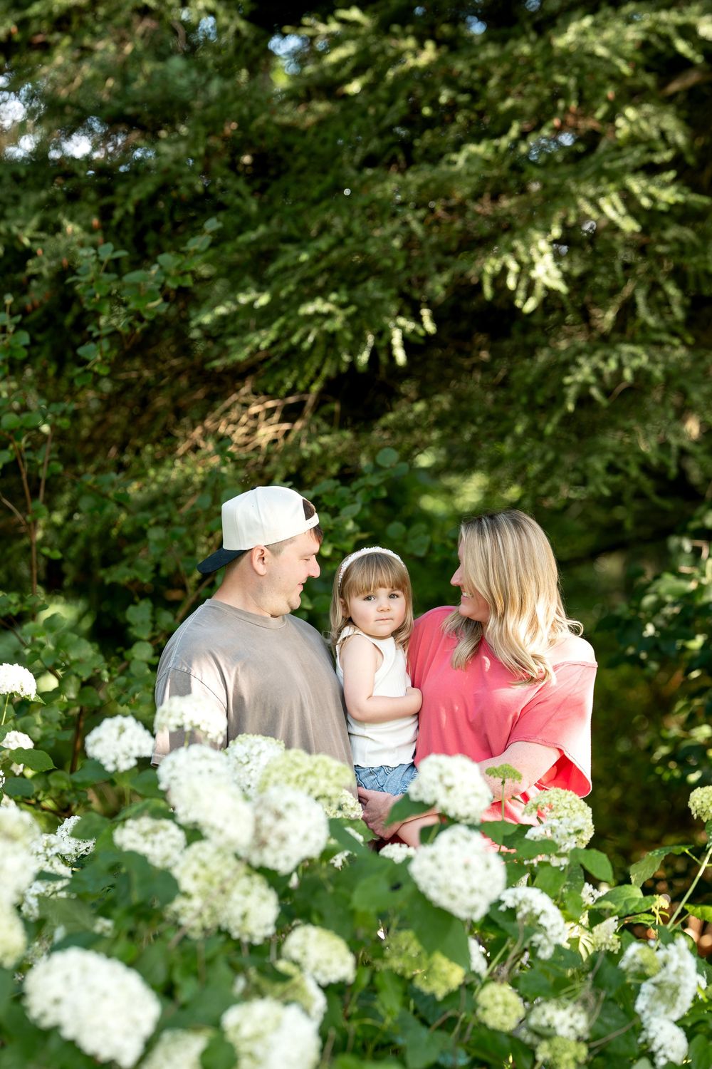A happy family with a toddler poses among white hydrangeas and lush green trees in a sunlit garden.