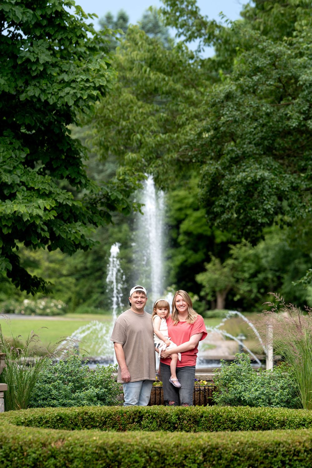 Family of three posing in a lush garden with a fountain backdrop on a sunny day.