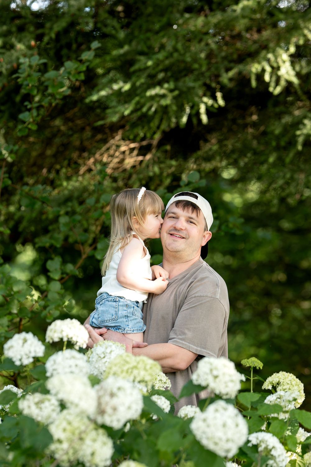 Little girl kisses man in gray shirt and cap while surrounded by white hydrangea flowers.