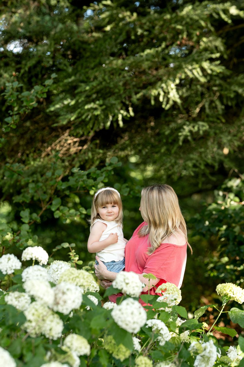 Mother holding her young daughter among white hydrangea flowers in a green garden.