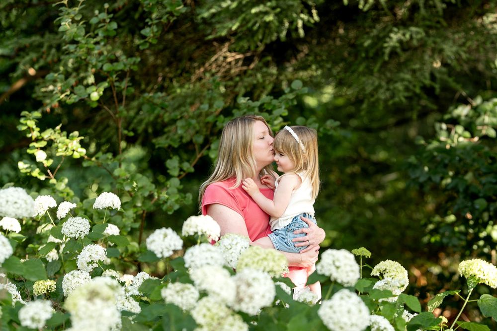 Mother kissing young daughter surrounded by white hydrangea flowers in lush garden.
