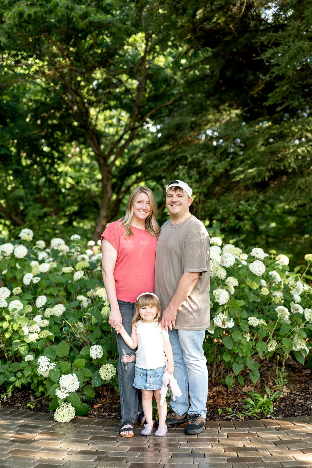 Happy family of three posing together in front of lush white hydrangea bushes in a green park setting.
