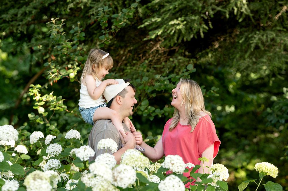 Family with young child on shoulders posing among white hydrangeas in lush green garden setting.
