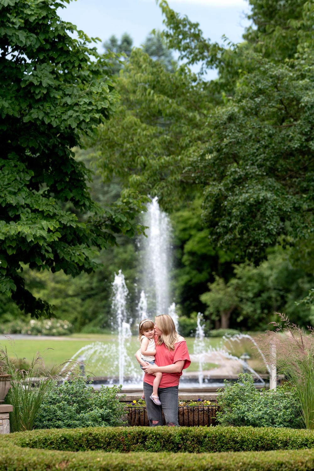Mother in red top cradles young child near ornamental garden with large water fountain backdrop.