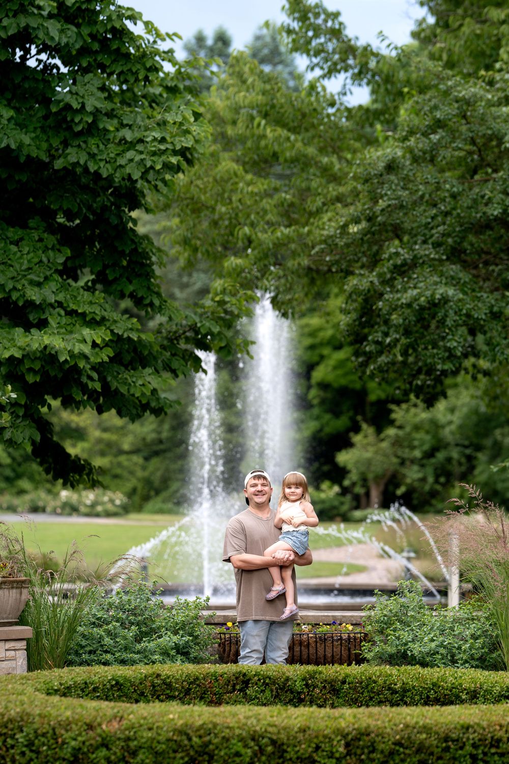 Father in tan shirt holds young toddler standing before a park fountain with green trees.