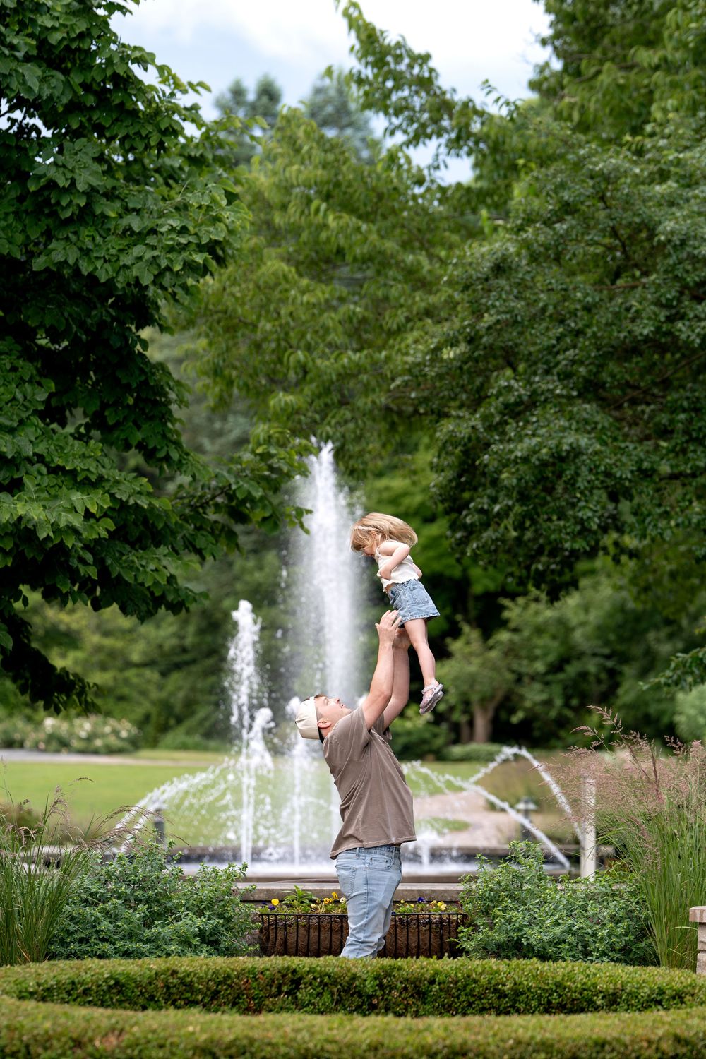 Man lifting toddler in the air in front of a fountain in a lush green park.
