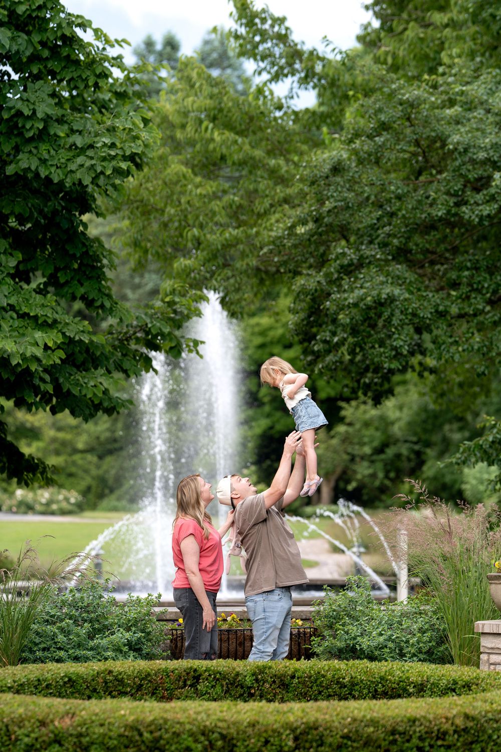 Family enjoying a sunny day at a park fountain, with father lifting young daughter in the air while mother watches.