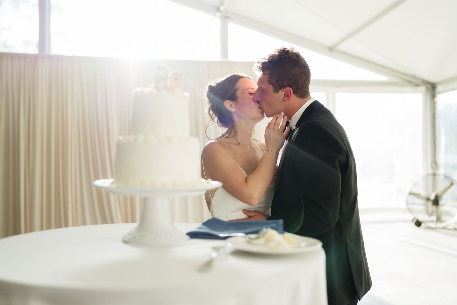 Bride and groom sharing a kiss beside a white wedding cake at their reception.